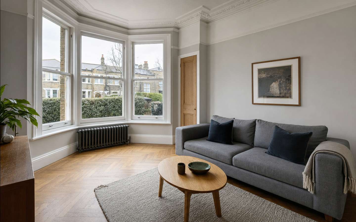 A living room in a Victorian terraced house: a bay window with white sash panes looking out to brick terraces, cornicing, a cast-iron radiator and a herringbone floor.